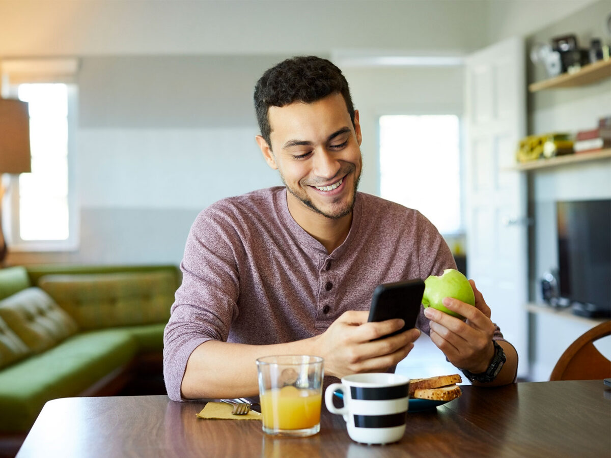 male smiling while looking at his phone and eating an apple for breakfast