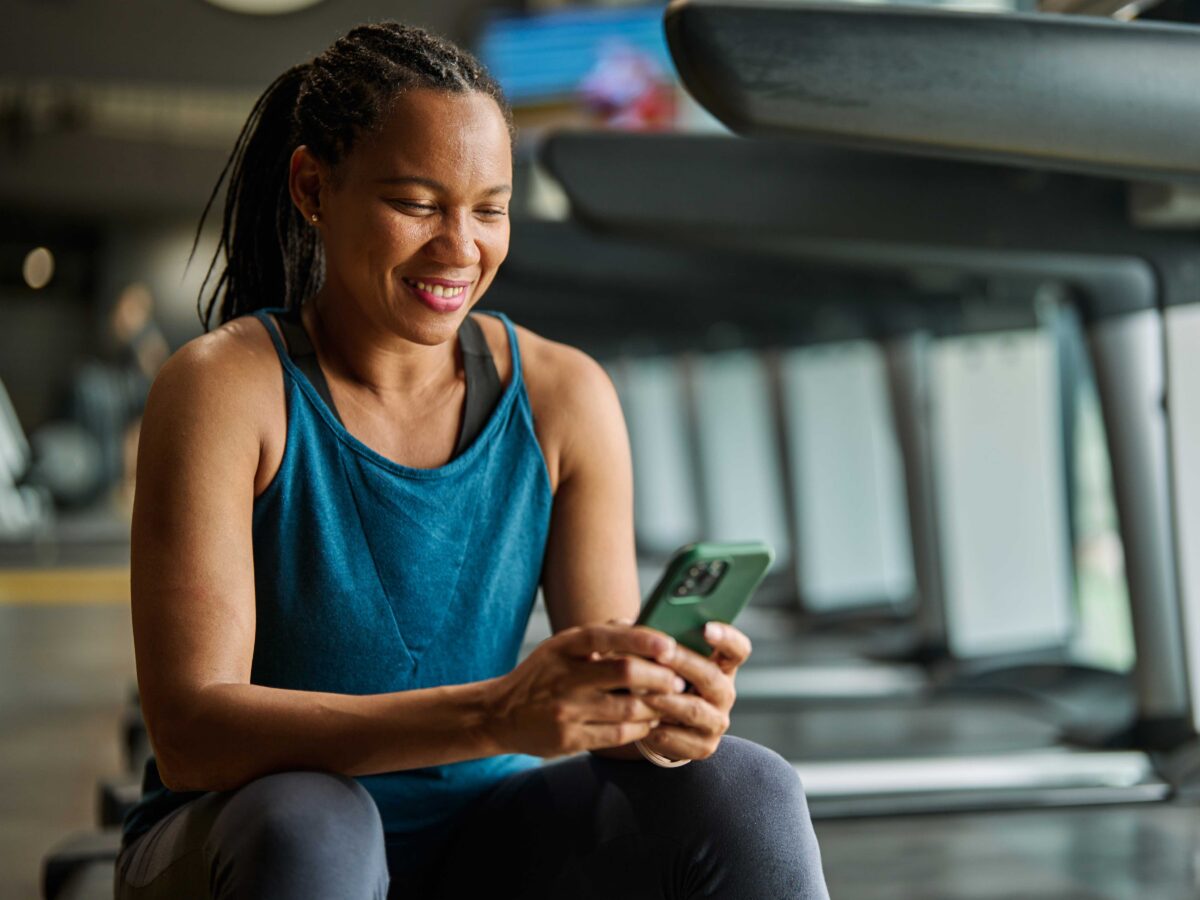 female smiling while sitting on the treadmill and looking at her wellness rewards and incentives on her phone